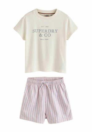 Młoda dziewczyna z długimi blond włosami, ubrana w białą koszulkę Superdry, różowe paskowane spodenki, białe skarpety, brązowe buty, trzymająca kremowy sweter z dzianiny.