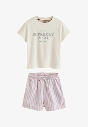 Ung pige med langt blondt hår iført en hvid Superdry t-shirt, stribede pink shorts, hvide sokker, brune sko og holder en cremefarvet strikket sweater.