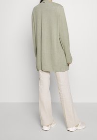 Cardigan vert clair à manches longues avec une coupe décontractée, bords côtelés et fentes sur les côtés, associé à un pantalon large beige clair et des baskets blanches.