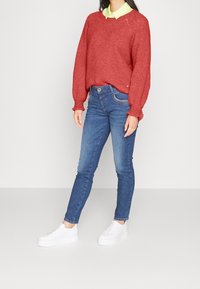 Rode gebreide trui, ontspannen pasvorm met ribbels aan de mouwen, gecombineerd met slim-fit blauwe jeans en witte platform sneakers. Groene overhemd met kraag eronder.