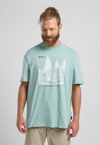 Hellblaue Baumwoll-T-Shirt mit einem weißen Grafikdesign von Bäumen und einem Zelt, mit dem Text "Platz zum Herumreisen schaffen" unter der Illustration.