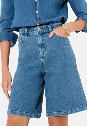 Persoană purtând pantaloni scurți din denim albastru de lungime medie și o cămașă de denim albastru cu mâneci suflecate, cu mâna odihnindu-se în buzunarul pantalonilor scurți.