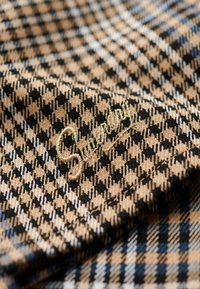 Tissu pied-de-poule marron et noir avec un logo brodé en or. La texture est douce avec un motif tissé sur toute la surface.