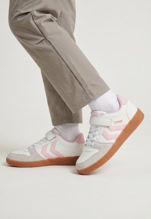 White and pink sneakers with gum soles worn with white socks and beige pants, one foot slightly raised against a white background.