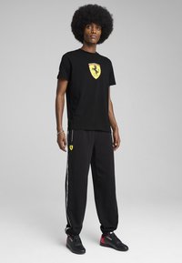 Čierne bavlnené tričko s žltým logom Ferrari, v kombinácii s čiernymi teplákmi so bočnými pruhmi a červenými akcentmi na topánkach.