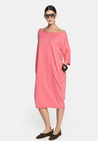 Luźna sukienka bez ramiączek w intensywnym różowym kolorze z bawełny, z krótkimi rękawami i kieszeniami bocznymi. Noszona z ciemnymi butami wsuwanymi i dużymi okularami przeciwsłonecznymi.