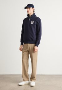 Pulover în nuanță de bleumarin cu închidere prin jumătate de fermoar și logo pe piept, asortat cu pantaloni khaki și adidași albi. Modelul poartă o șapcă bleumarin peste o cămașă în carouri.