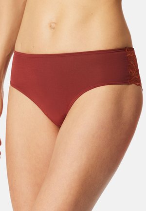 Frau trägt hipster Slips in Rostrot mit Spitzendetails an den Seiten, zeigt Mittelteil und obere Oberschenkel vor hellem, einfarbigem Hintergrund.