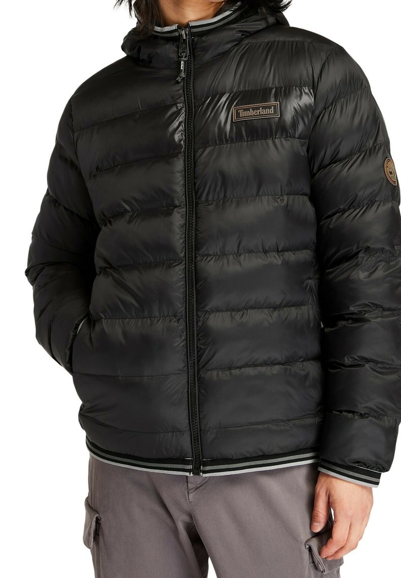 Personne portant une doudoune noire Timberland avec capuche, fermeture éclair à l'avant, poignets rayés et pantalon cargo marron.