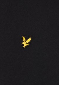 Tela negra con punto texturizado; presenta un pequeño emblema bordado de un pájaro amarillo en el área del pecho izquierdo. Diseño simple y limpio.