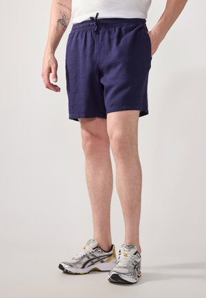 Shorts en lin bleu marine avec une taille élastique et un cordon de serrage. Portés avec des chaussures de sport grises et jaunes. Comprend des poches latérales.