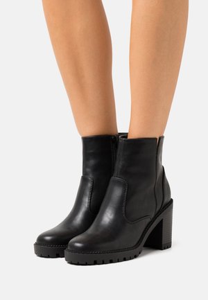 Stivali alla caviglia in pelle nera con tacco spesso e cerniere laterali indossati su gambe nude su uno sfondo bianco.