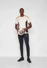 Nike spordiriietus, millel on valge ja must lühikeste varrukatega topp kuldsete aktsentidega, koos mustade pükstega. Mängija hoiab käes mustritest jalgpalli.