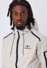 Giubbotto a vento beige chiaro con accenti neri, zip frontale, cappuccio regolabile e logo. Presenta una texture liscia e una vestibilità atletica.