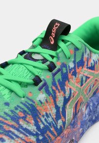 Zapatilla deportiva verde con un upper texturizado, que presenta patrones abstractos coloridos en azul y naranja. Incluye cordones negros y una etiqueta con el logo.