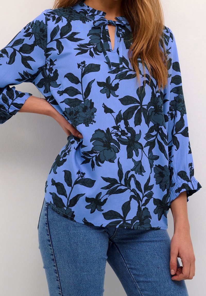 Femme portant un chemisier bleu à imprimé floral foncé avec des manches trois-quarts, associé à un jean bleu, debout contre un mur clair.
