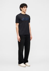 Νέος άνδρας που στέκεται, φοράει μαύρο t-shirt Armani Exchange, μαύρο τζιν και λευκά αθλητικά παπούτσια, με φόντο ένα απλό ανοιχτό γκρι τοίχο.