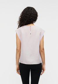 Ανοιχτό μωβ μπλούζα με κοντά μανίκια, λεία υφή, ψηλός λαιμός και μοναδική λεπτομέρεια με κουμπί στο πίσω μέρος. Συνδυάζεται με σκούρο παντελόνι.