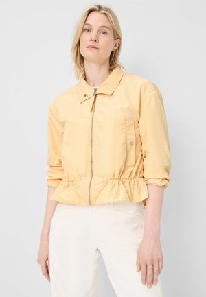 Blonde Frau, die eine hellgelbe Jacke mit Reißverschluss und geraffter Taille sowie eine weiße Hose trägt, steht vor einem schlichten hellgrauen Hintergrund.