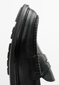 Zwart leren schoen met een ronde neus, voorzien van uitgesproken stiksels en een klittenbandsluiting. Dikke zwarte rubberen zool met grip.