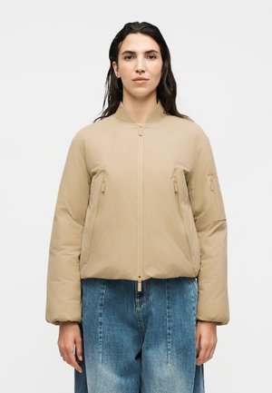 Femme aux longs cheveux foncés portant une veste matelassée beige zippée et un jean bleu ample, debout devant un fond blanc uni.