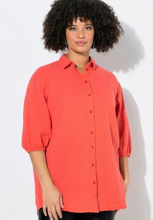Kvinde med krøllet hår iført en løstsiddende, koralfarvet skjorte med knapper og trekvartlange ærmer, stående mod en hvid baggrund.
