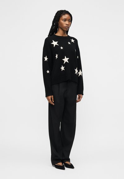 Donna che indossa un maglione nero con stelle bianche, pantaloni neri larghi e scarpe nere a punta, in piedi contro uno sfondo bianco semplice.