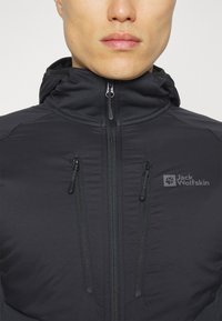 Black, hooded jacket with two zippered pockets. Made of textured fabric, featuring the "Jack Wolfskin" logo on the left chest area.