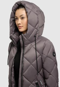 Quilted puffer jacket in muted gray with a large hood, black drawstrings, and zippered pockets. Smooth, shiny texture, featuring a logo patch.
