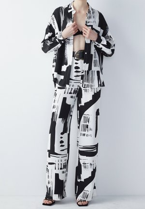 Person trägt ein schwarz-weiß abstrakt gemustertes, oversized Hemd und passende weite Hosen, schwarzer Gürtel und schwarze Sandalen, Hände richten das Hemd.