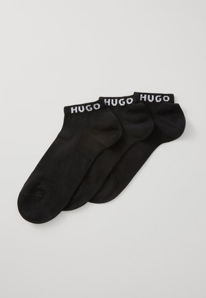 Tri para crnih čarapa do gležnja s natpisom "HUGO" u bijelim slovima na rubovima, preklopljenih jedna preko druge na svijetloj pozadini.