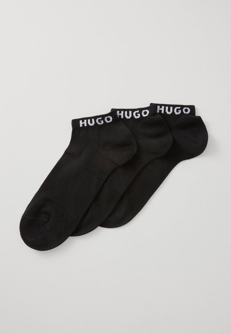 Tři páry černých kotníkových ponožek s bílými nápisy "HUGO" na lemu, položené překrývající se na světlém pozadí.