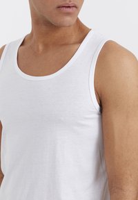 Weißes Baumwoll-Top mit rundem Halsausschnitt und dünnen Trägern, das eine glatte Textur und ein einfaches, unaufdringliches Design aufweist.