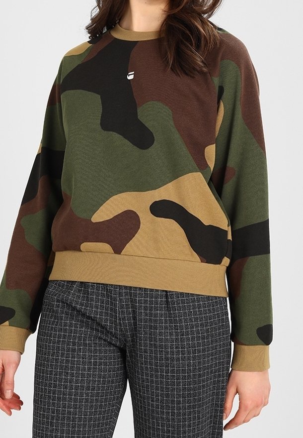 Woman wearing a green, brown, and black camouflage sweater with beige cuffs and grey plaid pants against a plain background.