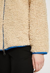 Beige fleecová bunda s texturovaným, kudrnatým materiálem; modré zip a lemování; viditelný tahák na zip; nošena přes černé kalhoty.