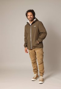 Bruine waterdichte jas met een fleece voering, witte rits en elastische manchetten, gedragen over beige cargo broeken en tan sneakers.