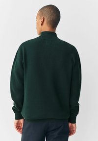 Hombre con cabello corto, vistiendo un suéter de punto acanalado en verde oscuro y pantalones oscuros, de pie mirando hacia atrás contra un fondo claro y liso.