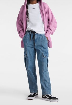 Persona que lleva una sudadera con capucha rosa, camiseta blanca de Vans, vaqueros cargo azules con cordón negro y zapatillas Vans blancas y negras, de pie con las manos en los bolsillos.