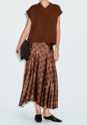 Femme portant un haut sans manches en tricot marron, une jupe en satin marron à carreaux, des chaussures noires sans lacets et tenant une pochette sombre.