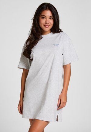 Junge Frau mit langen dunklen Haaren, die ein lockeres, hellgraues Kurzarmkleid mit kleinem blauen Text nahe der Brust trägt.