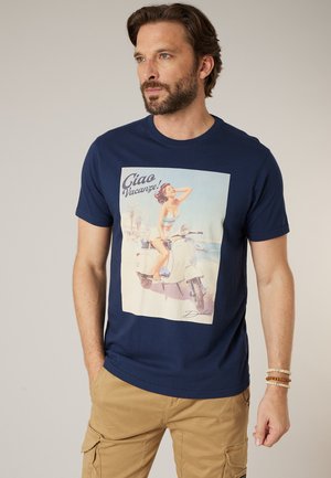Man with beard wearing navy blue t-shirt featuring a vintage-style print of a woman on a scooter by the beach, paired with tan cargo pants.