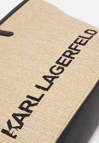 Béžová tkaná taška přes rameno s černým vyšívaným nápisem "KARL LAGERFELD", vybavená černým koženým dnem a horním uchycením.