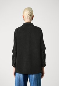Black corduroy shirt jacket with a back yoke, long sleeves, buttoned cuffs, and a straight hem. Worn over light blue denim.