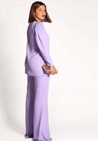 Conjunto de dos piezas en lila que incluye una blusa de manga larga con hombros abullonados y pantalones de pierna ancha. Accesorios en dorado con un clutch. Textura suave.