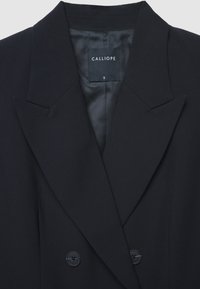 Blazer nero doppiopetto con colletto a revers, due grandi bottoni neri e una fodera liscia in satin con etichetta all'interno.