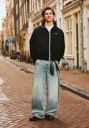 Young man in black zip-up jacket and wide-leg jeans standing on a brick street lined with bicycles and traditional European buildings.