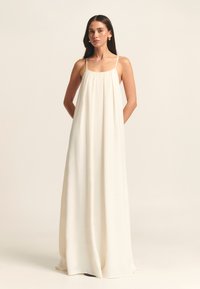Robe maxi à fines bretelles, confectionnée en tissu léger de couleur crème. Présente un design ample et fluide avec un buste froncé et sans motifs visibles.