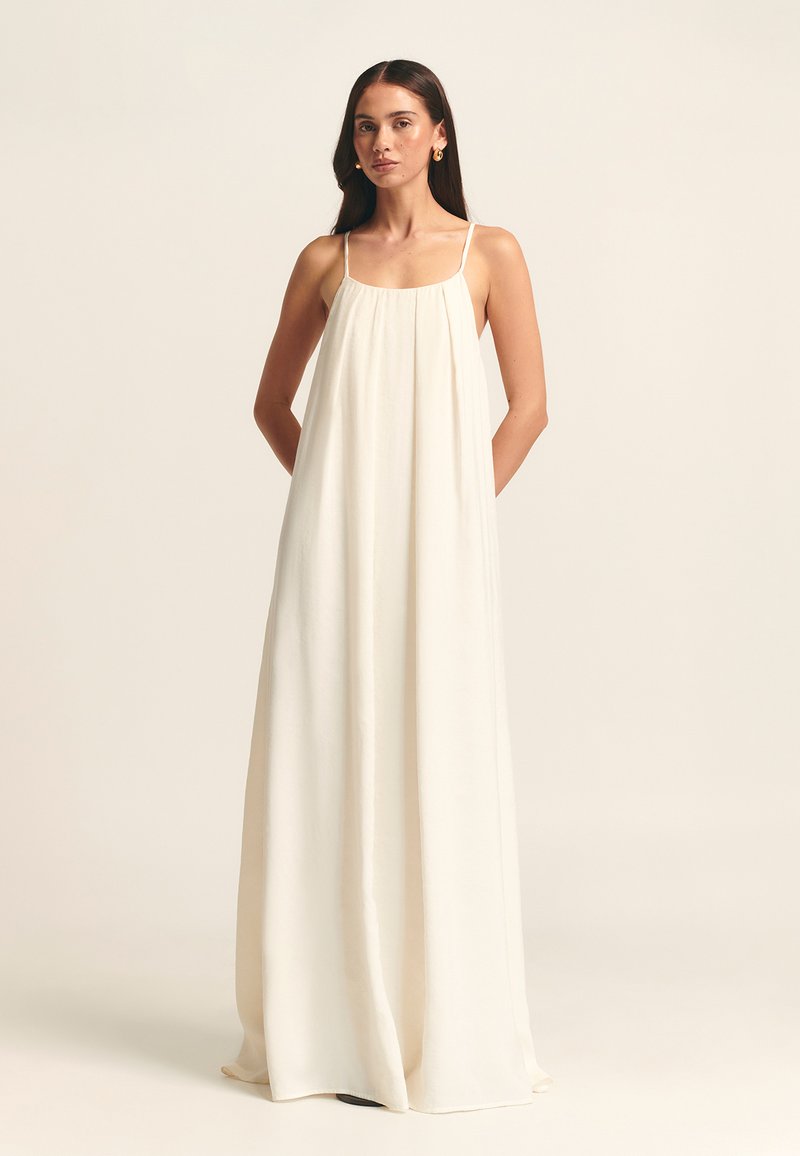 Robe maxi à fines bretelles, confectionnée en tissu léger de couleur crème. Présente un design ample et fluide avec un buste froncé et sans motifs visibles.