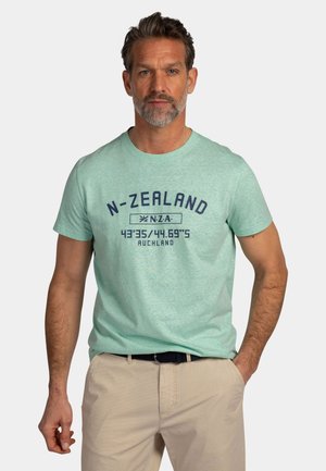 Man met kort bruin haar en baard, een lichtgroen T-shirt met Nieuw-Zeelandse afbeelding en beige broek, staand met één hand in de zak.