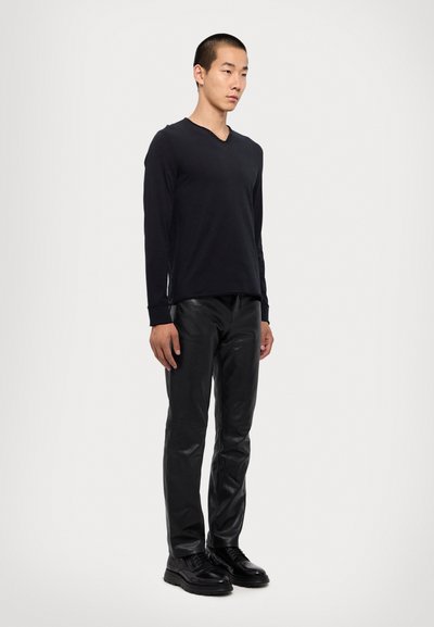 Chemise noire à manches longues en V, pantalon en cuir noir et chaussures à lacets noires portés par un mannequin debout sur un fond blanc uni.